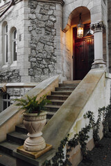 Obraz premium The architectural structure is built of stone in gray color, with window, decorated with green plants. Concrete stairs in the foreground. An antique brown door illuminated by a lantern.