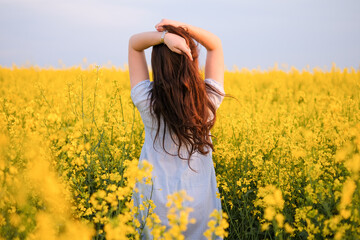 Woman in rapeseed field blooming in the spring. Back view of girl with long brown hair enjoying sun in blossom of canola. Freedom concept.