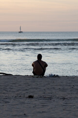man sitting on sand by ocean watching sailboat at sunset
