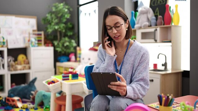 Young beautiful hispanic woman working as a teacher talking on smartphone using touchpad at kindergarten