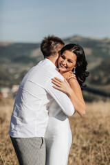 A stylish model couple in the mountains in the summer. A young boy and a girl in a white silk dress are walking on the slope against the background of the forest and mountain peaks. boy kisses a girl