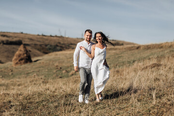 A stylish model couple in the mountains in the summer. A young boy and a girl in a white silk dress are walking on the slope against the background of the forest and mountain peaks. boy kisses a girl