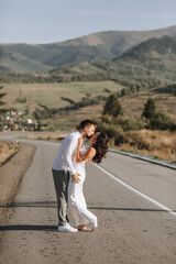 Fototapeta premium A stylish groom in a white shirt and a cute brunette bride in a white dress are hugging and kissing on an asphalt road against the background of a forest and mountains. Wedding portrait of newlyweds.