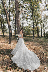 Wedding photo in nature. The bride is standing in the forest. The bride in a beautiful dress with a long train, holding her bouquet of white roses, smiling sincerely at the camera. Portrait