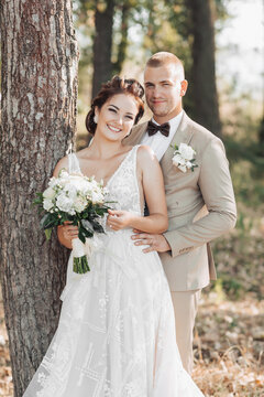 Wedding photo in nature. The bride and groom are standing near a tree smiling and looking at the camera. The groom hugs his beloved from behind, the bride holds a bouquet. Portrait. Summer wedding
