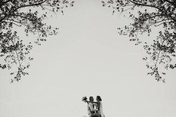 Atmospheric wide-angle photo against the background of the sky and trees. The bride, holding a bouquet, hugs the groom. Black and white photo. Wedding portrait