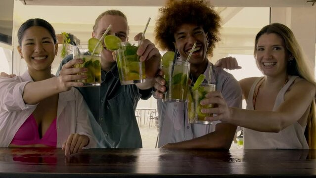 Multiracial friends toasting with mojito cocktails at beach bar looking at camera. 4k video of friends having fun.