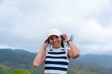 Mujer latina sonriendo feliz con el cabello movido por el viento, viendo a cámara con una montaña...