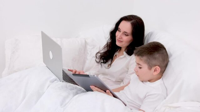 mom and son under white blanket are sitting on soft bed in the hands of boy tablet playing social networks online he is 4-6 years old woman works as freelancer on laptop remote work online conference