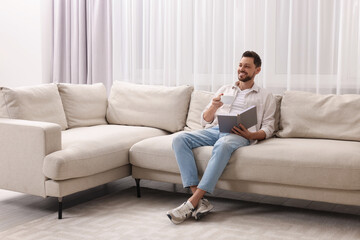 Fototapeta na wymiar Happy man drinking coffee while reading book on sofa near window with beautiful curtains at home in living room. Space for text