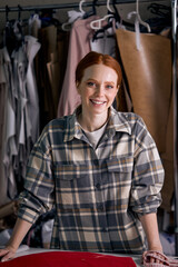 excited caucasian dressmaker woman sews clothes at tailor office, engaged in tailoring,redhead lady in checkered shirt posing at camera, fabric clothes in the background. dressmaking industry
