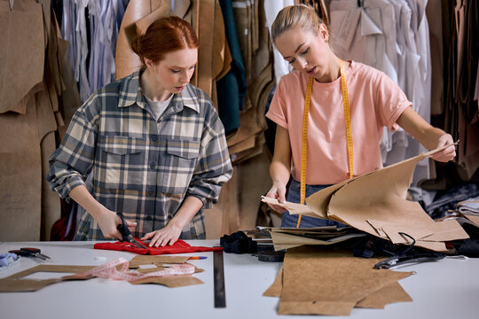 two young caucasian hardworking seamstresses in modern studio workshop, preparing textile to sew, redhead and blonde ladies in casual wear in fabric, having talk, clothing manufactory production