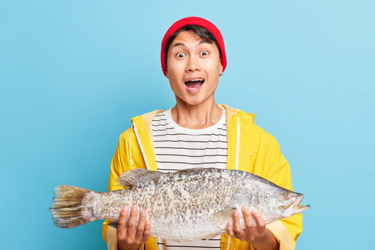 Happy guy dressed in red cap and yellow jacket holding big fish in his hands, showing enjoyment by such a good catch, fortune fishing concept, copy space, high quality photo