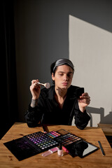 glamour man blowing kiss, holding mirror and brush sitting at the table at home. close up portrait, cosmetics. lifestyle, hobby, interest