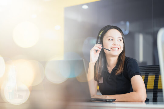 Friendly Asian female officer smiling happily in headset working on computer in call center.