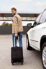 Back view, serious man walking with suitcase, holding smartphone, near car. Concept of travel, road, car trip