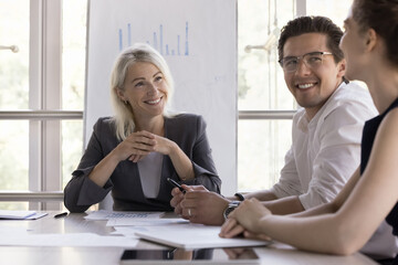 Happy middle aged female leader listening to young employees ideas, offers on brainstorming...