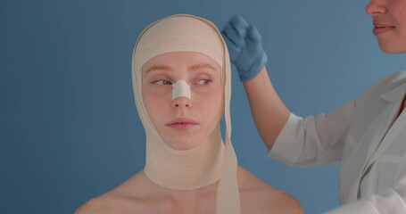 Doctor cover the new face of female patient by bandage isolated on a blue background, closeup portrait. Plastic Surgeon bandaging head of young beautiful woman. Beautiful Girl after Plastic Surgery