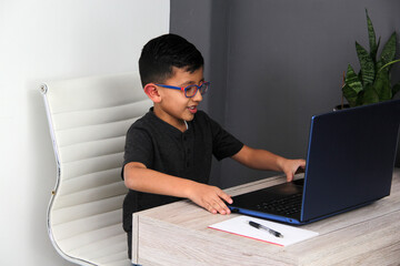 7-year-old Latino boy with glasses does home schooling takes online classes at home on a desk with a laptop, studies, is surprised and participates in class