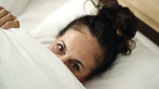 Middle age hispanic woman lying on bed covering face with bedsheet at bedroom