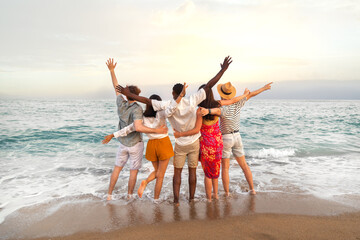 Rear view of multiracial friends embracing together looking at the ocean celebrating with arms up...
