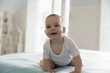 Fototapeta premium Curious positive few month baby crawling on knees, looking away, smiling, laughing, feeling joy. Infant boy or girl in white bodysuit moving on soft clean mattress. Innocent little kid home portrait