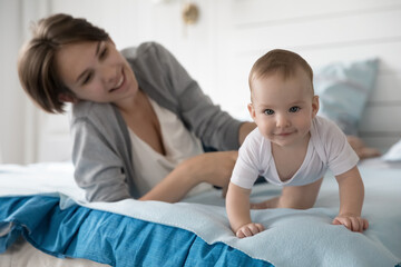 Obraz na płótnie Canvas Positive adorable baby crawling on knees from happy mom to camera, moving on bed, resting on clean linen with laughing young new mother. Mum cuddling kid in home bedroom, enjoying motherhood