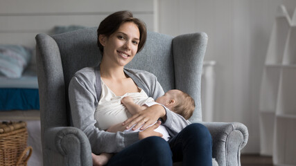 Obraz premium Happy beautiful young new mother breastfeeding infant child, resting in comfortable armchair at home, holding sleepy eating baby in arms, looking at camera, smiling, posing. Banner shot