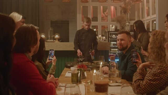 Male chef prepares meal in flambe style in front of people in modern gastro cafe. They film the process on their phones. Group of friends spend weekend evening in restaurant. Concept of public eating.