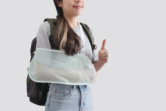 Travel insurance concepts. Young woman with hand injured wearing splint, broken arm, wearing backpack, showing thumb up, smiling face, isolated on white background - Powered by Adobe
