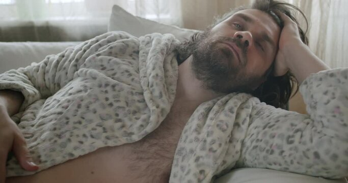 Carefree, careless lazy beard man in bathrobe lying on the couch scratching head. Unkept, shabby, pensive look. Procrastination, waste time and motivation problem. Laziness and procrastination concept