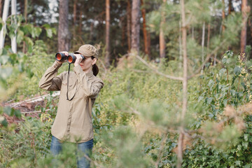 A woman forester in uniform looks through binoculars and monitoring the forest area in summer, selective focus.