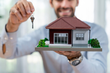 Focus house sample model and key held by blurred smiling and happy real estate agent or buyer in...