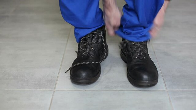 A worker in blue overalls puts on safety boots and ties his shoelaces. Safety engineering.
