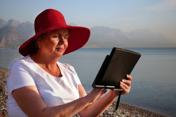 Senior woman holding tablet computer sitting on the beach, wearing in big red sunhat and taking sun bath