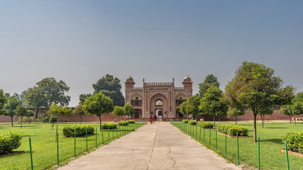 Fototapeta na wymiar The entrance to the ancient tomb of Itmad-Ud-Daulah. The red sandstone walls are decorated with white ornaments. People walk along the path through the arched gate. Grass and trees on the roadsides