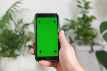 A man holds a phone with a green screen in his hands a background house plants. Mockup. Chromakey.