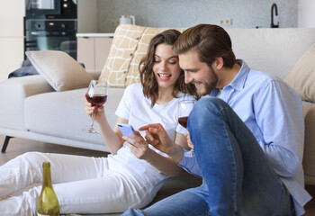 Happy couple sitting, relaxing on floor in living room, drinking red wine. Smiling young husband and wife rest at home enjoy romantic date on family weekend together