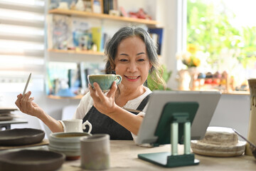 A retired potter woman using a tablet to sell pottery online, SME business ideas online. - obrazy, fototapety, plakaty