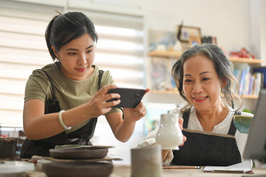 Pottery woman using smartphone to take pictures of pottery to sell online with her grandmother, family business, Art and craft concept.