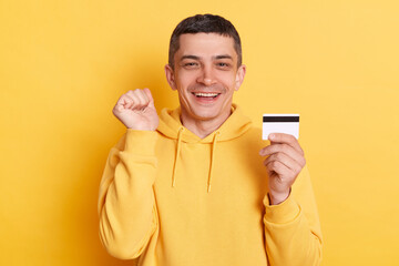 Online shopping. Banking paying. Delighted cheerful man wearing casual hoodie earning big sum of money clenched fist celebrating succes posing isolated over yellow background.