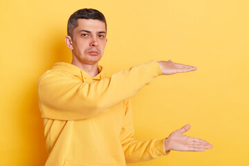 Sad disappointed man wearing casual hoodie showing big size empty area for advertisement looking at...