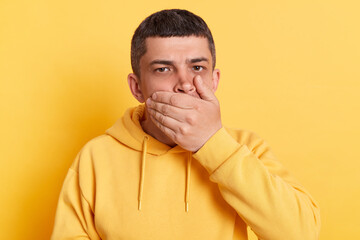 Shocked man wearing casual hoodie covering mouth with palm keeping secret looking at camera hearing...
