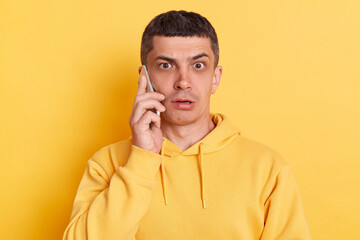 Shocked scared man wearing casual hoodie talking on smart phone hearing surprised news looking at camera with big eyes posing isolated over yellow background.