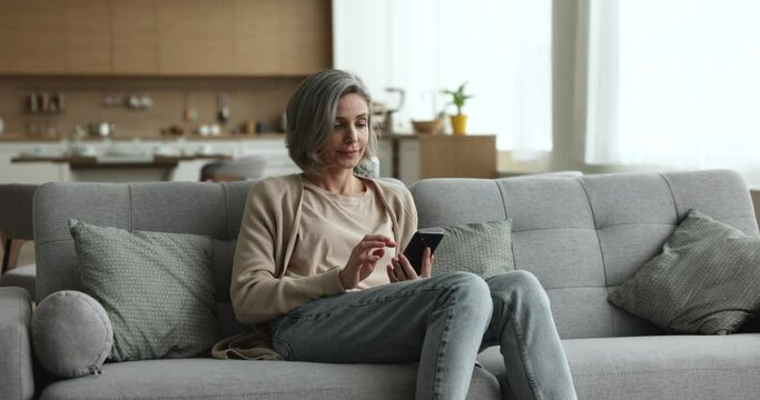 Focused grey haired woman using mobile app, online service on smartphone at home, shopping on webstores, enjoying Internet communication, leisure with digital gadget