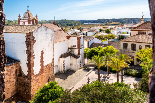 From castle of Silves, the Silves cathedral and its lateral square can be seen in front of Cidade de Silves rooftops, with palm trees and lush surroundings leading to Arade river under a sunny sky.