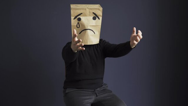 A sad man in a black turtleneck with a bag on his head, with a drawn crying emoticon, cries and asks for help stretching his hands forward. Emotions and gestures.