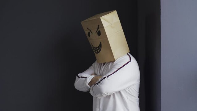 An angry man in a white shirt with a bag on his head, with an evil smiley face drawn, stands in the corner against the wall, laughs, waves his fists. Emotions and gestures.