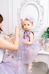 Fototapeta premium Baby girl elegant dress. A one-year-old girl in a puffy dress and a cute bow poses against the backdrop of a bright room with a dressing table and flowers.