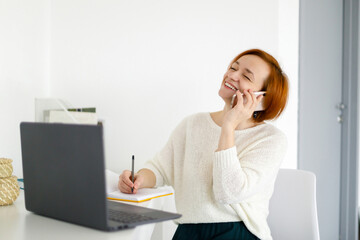 red haired woman in a white sweater sits at a white table and makes a plan for the day in a notebook, talking on the phone with a client. woman in business